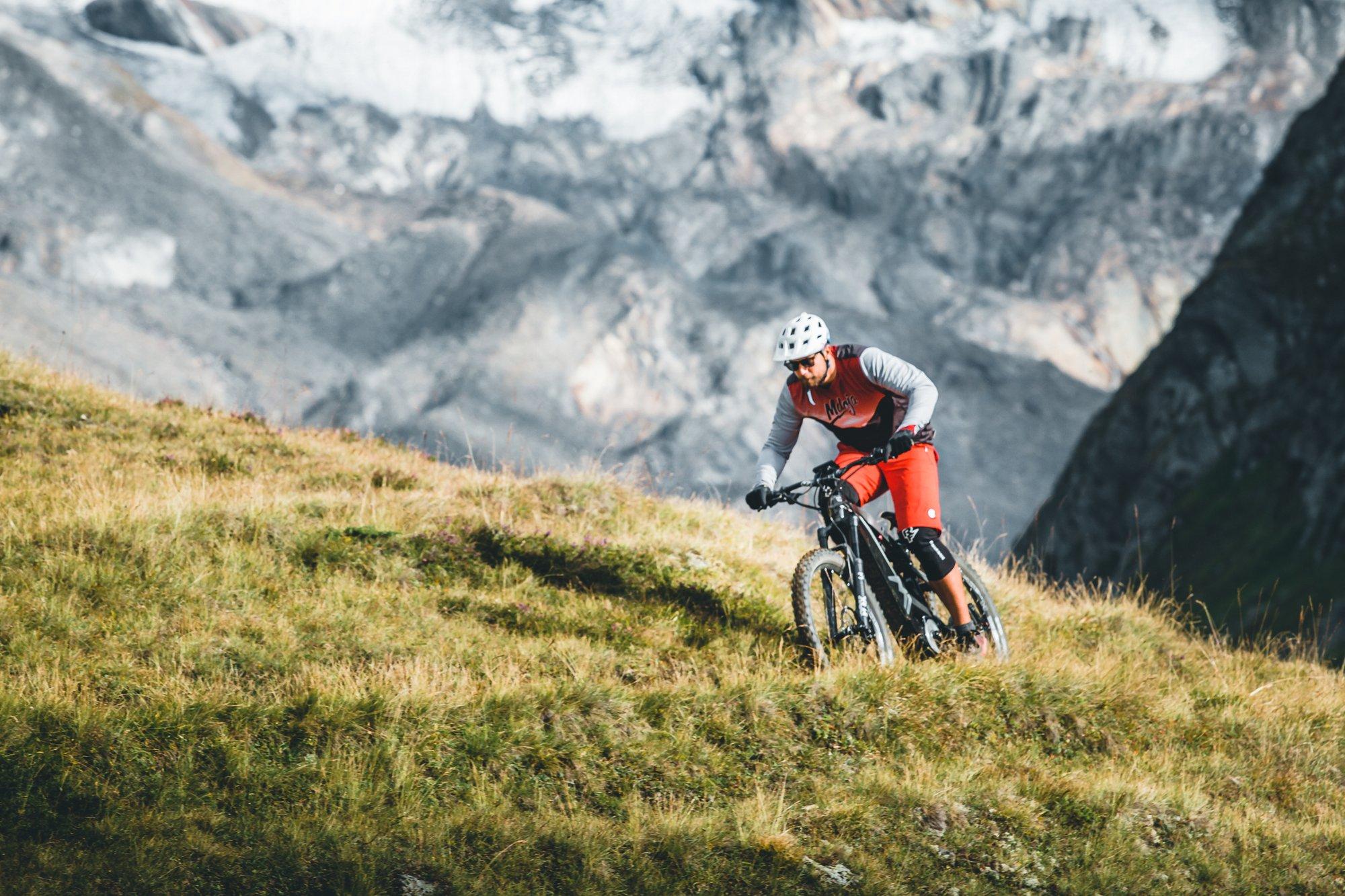 Ein Radfahrer auf einem Mountainbike fährt einen grasbewachsenen Hügel in einer alpinen Umgebung hinunter. Er trägt