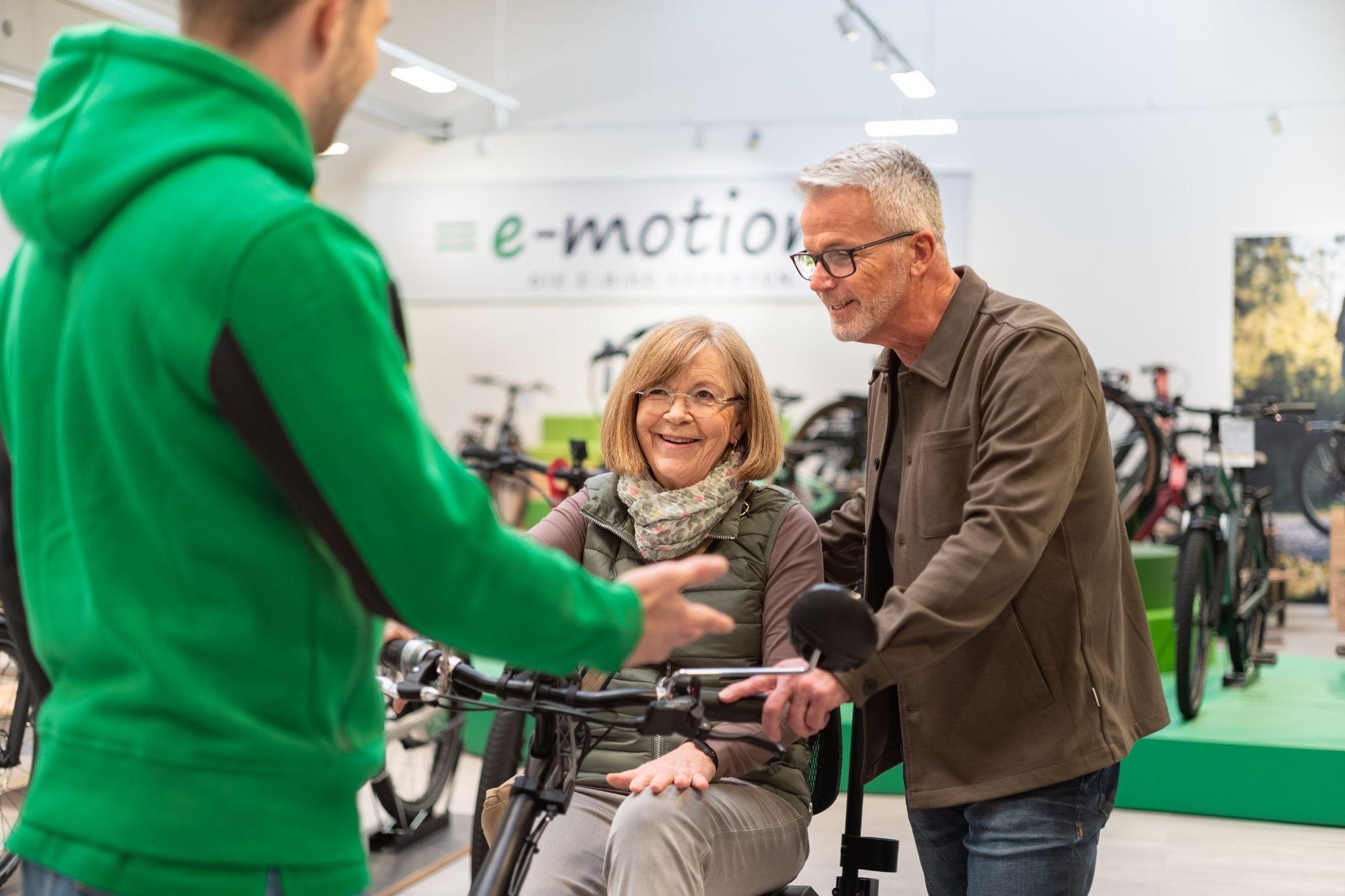 Eine Frau sitzt auf dem Dreirad, daneben steht ihre Begleitung und ein e-motion Berater