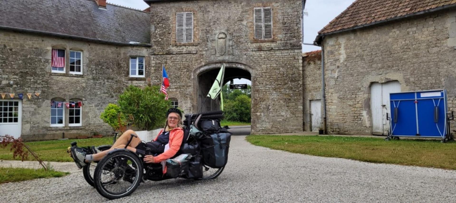 Eine Person sitzt auf einem beladenen Liegedreirad vor einem alten Steingebäude mit Torbogen und Fahnen.