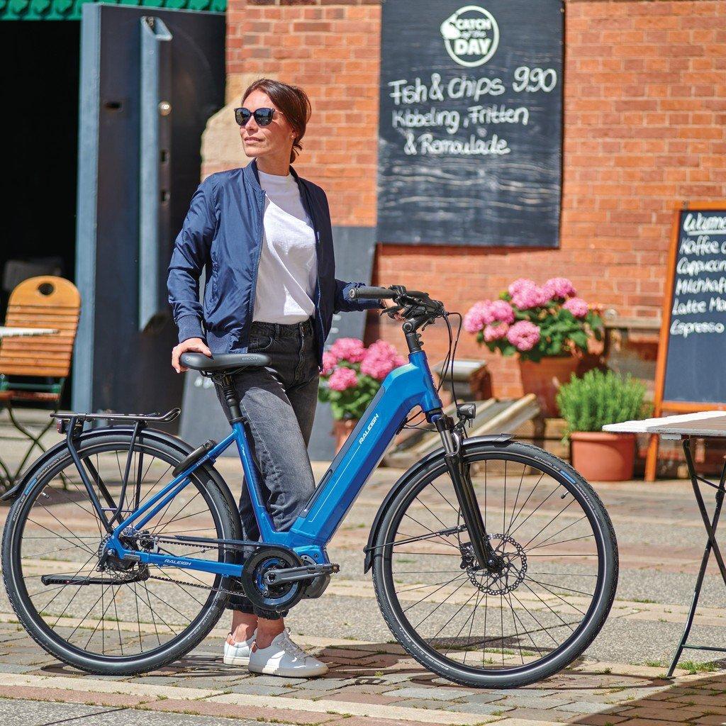 Eine Frau steht neben einem blauen E-Bike, das auf einem gepflasterten Platz vor einem Café parkt. Sie trägt Sonnen