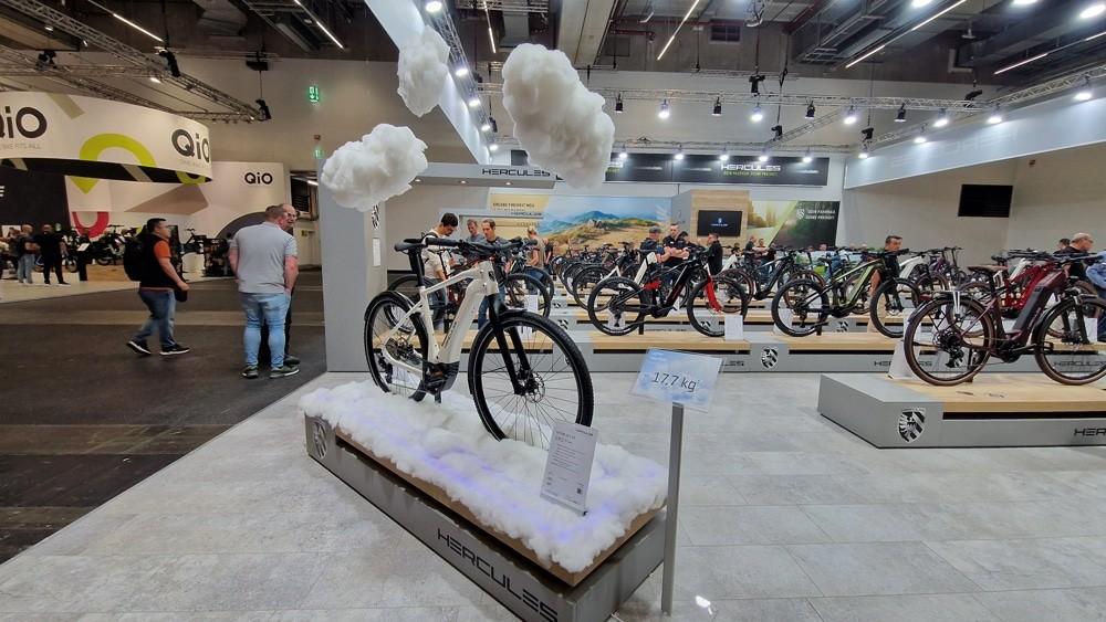 Hercules e-Bike auf Podest mit Wolkenkulisse, moderne Ausstellung mit weiteren Rädern und interessierten Besuchern.