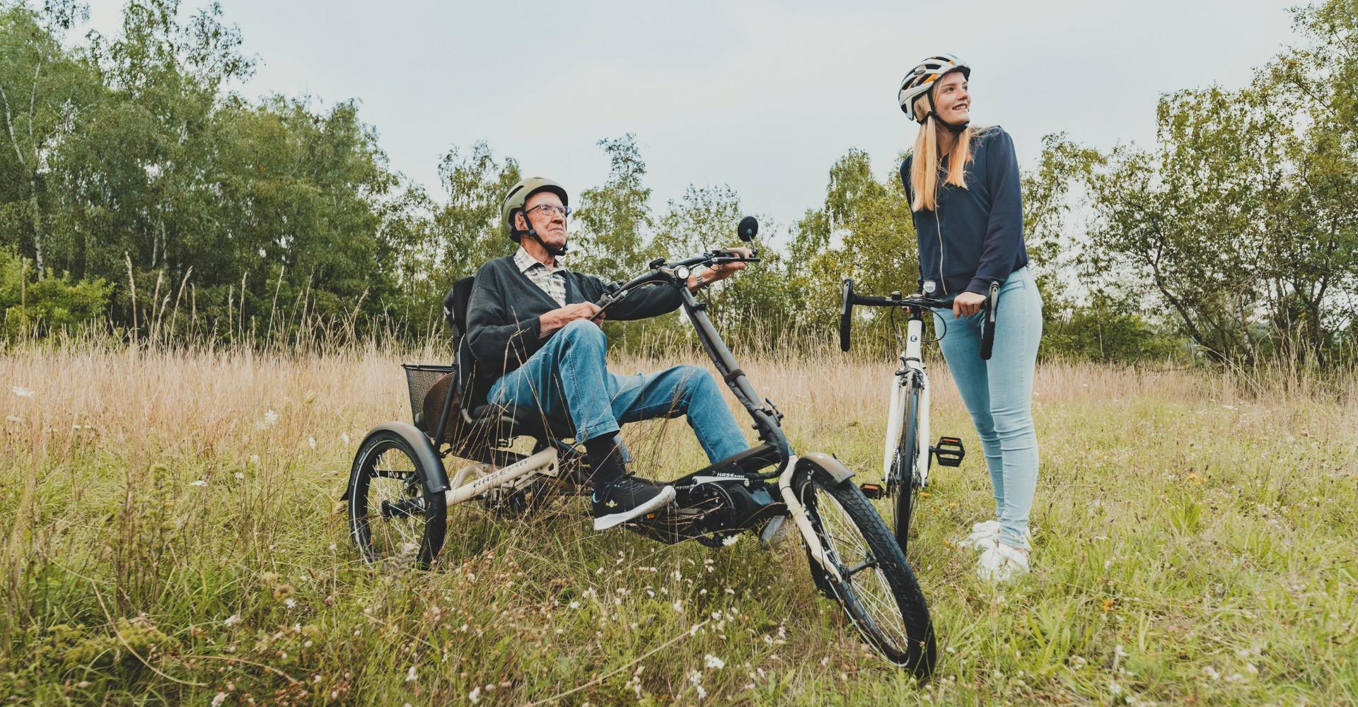 Ein Mann sitzt auf einem hase bikes Dreirad, neben ihm steht eine Frau mit einem herkömmlichen Fahrrad