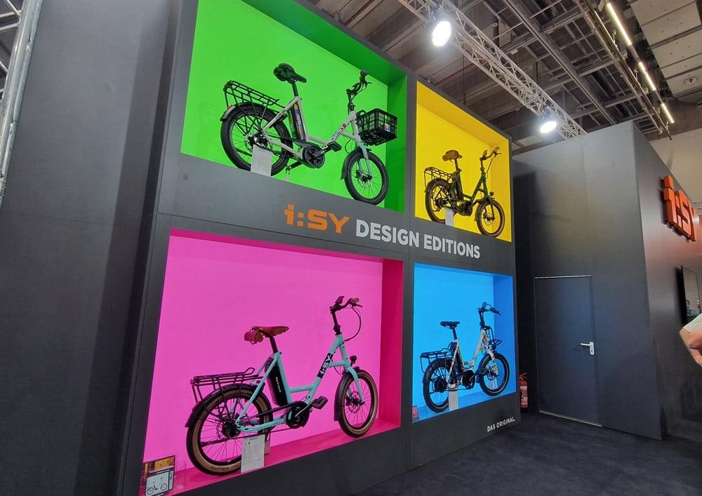 i:SY e-Bikes in bunten Kästen (grün, pink, gelb, blau) mit Gepäckträgern, moderne Ausstellung im kreativen Stand.