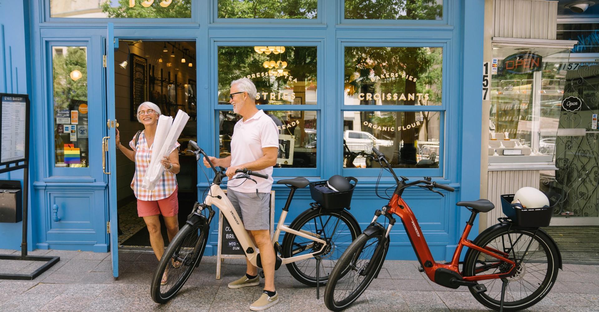 Eine Frau kommt aus einer Bäckerei, ein Mann wartet davor mit zwei Specialized e-Bikes