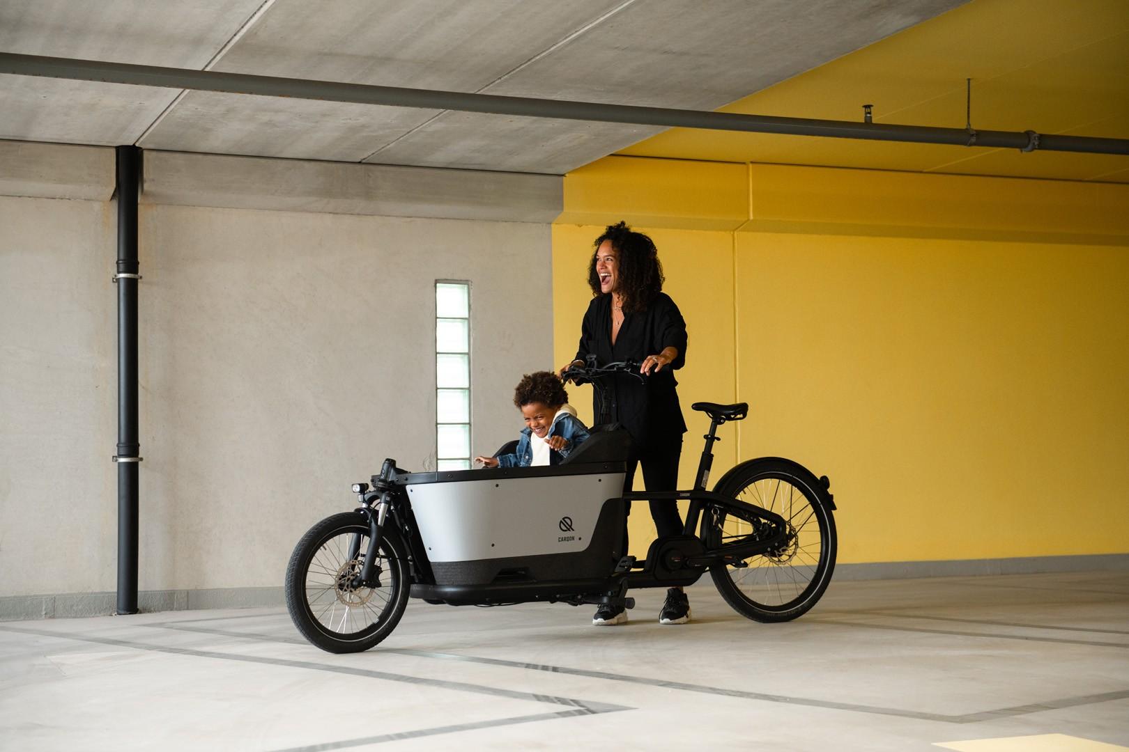 Carqon Cruise, eine Frau steht mit einem Kind in der Transportbox vor gelber Wand