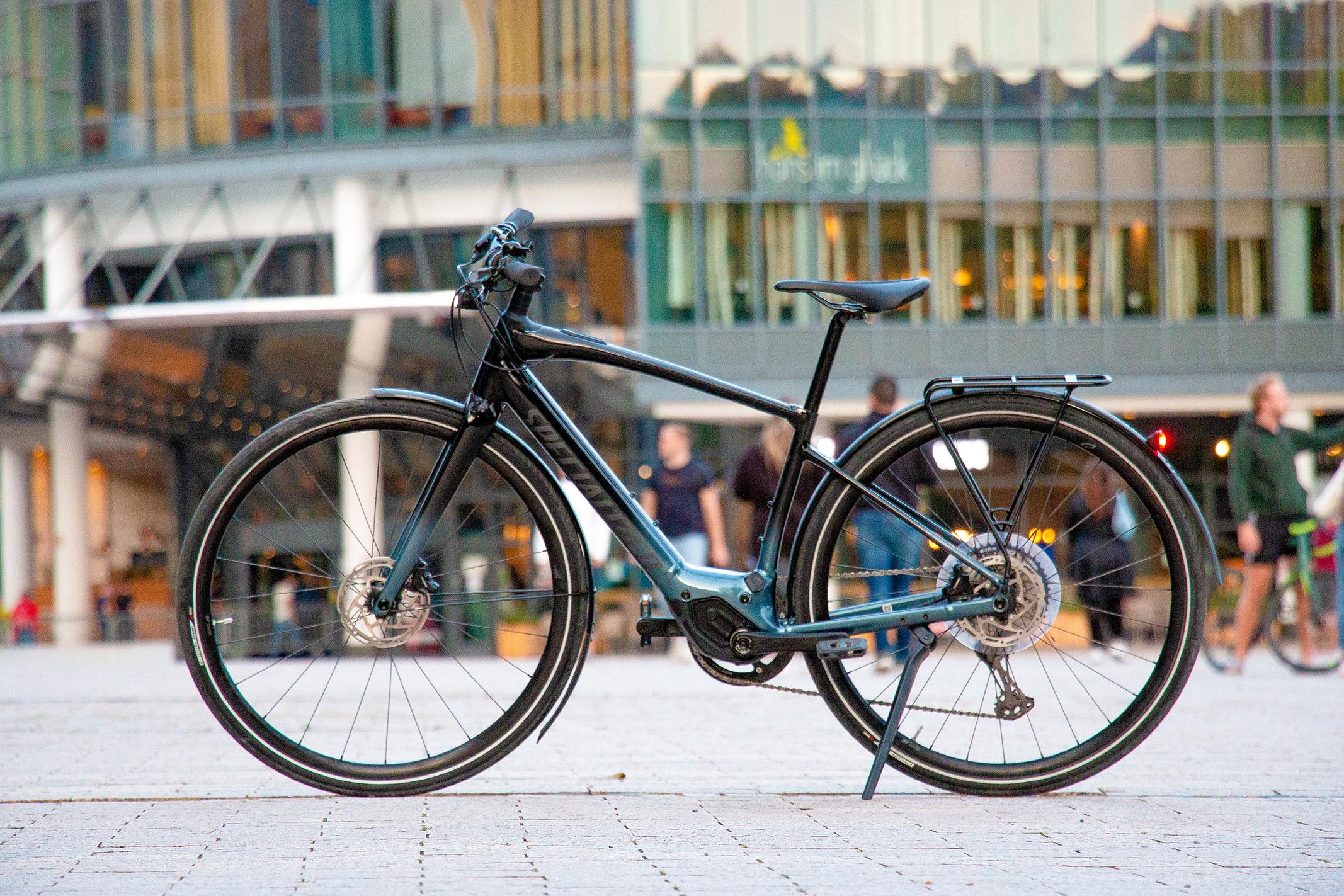 Specialized Vado SL E-Bike in Schwarz, seitlich auf einem Platz vor moderner Glasfassade aufgenommen.