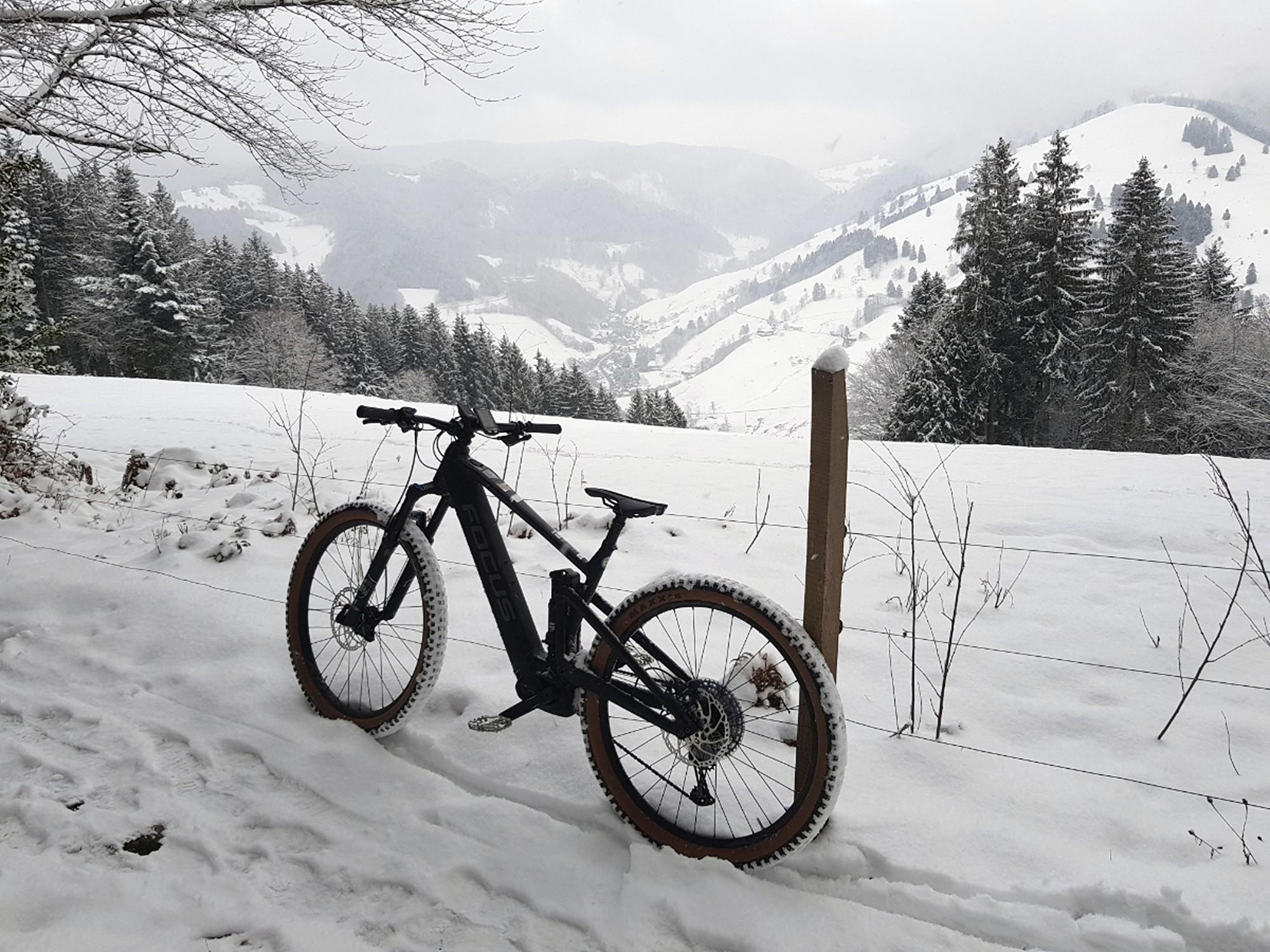 Das Bild zeigt ein E-Bike, das im Schnee eines malerischen Berglands geparkt ist.