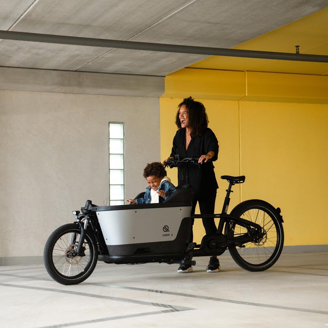 Carqon Cruise, eine Frau steht mit einem Kind in der Transportbox vor gelber Wand