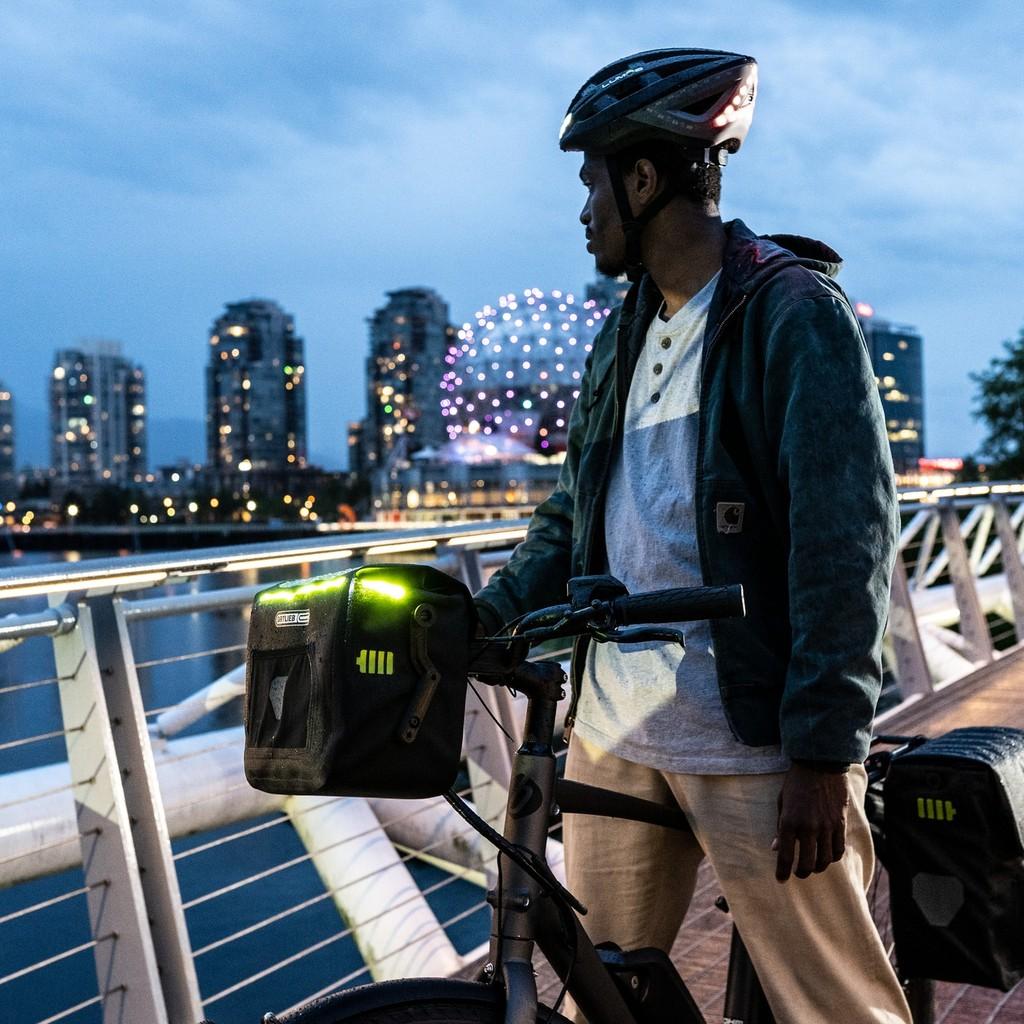 Mann auf einer Brücke mit einem VAN E-Bike und ORTLIEB Fahrradtaschen, vor einer städtischen Skyline.