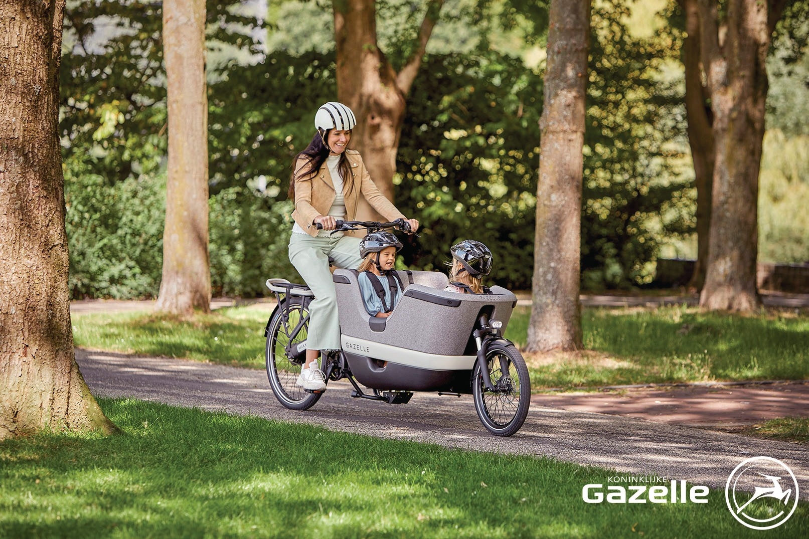 Eine Frau fährt das Gazelle Makki Load, in der Transportbox zwei Kinder mit Helm