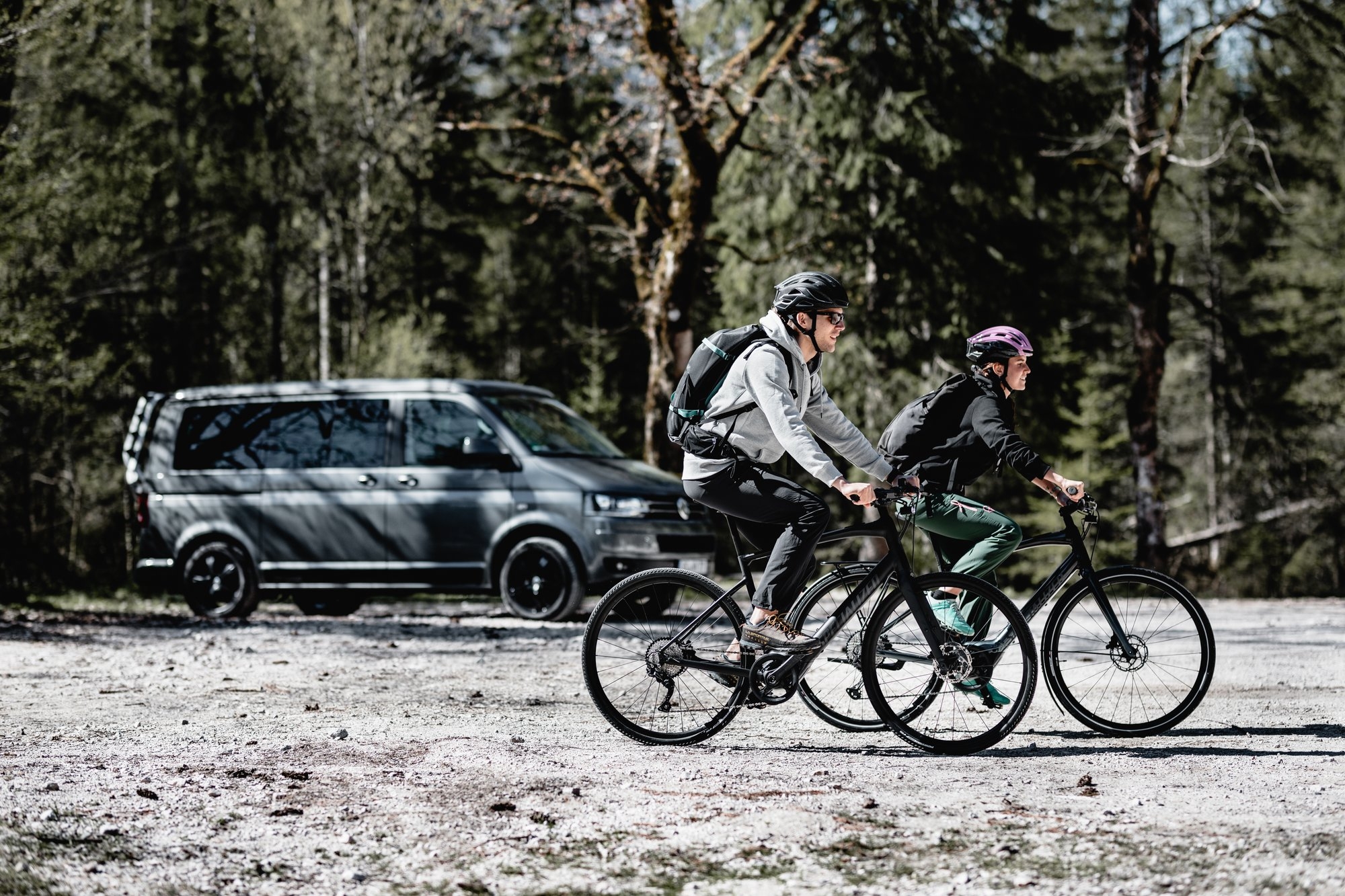Zwei Radfahrer tragen Helme und fahren nebeneinander auf einem Fahrradweg durch einen Wald. Im Hintergrund steht ei