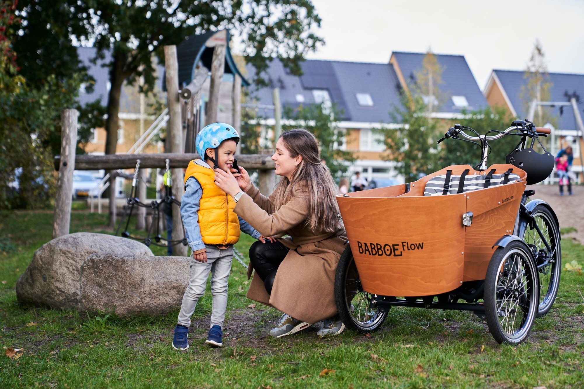 Eine Frau zieht einem Helm einem Kind an, daneben steht ein Babboe Flow Lastenrad