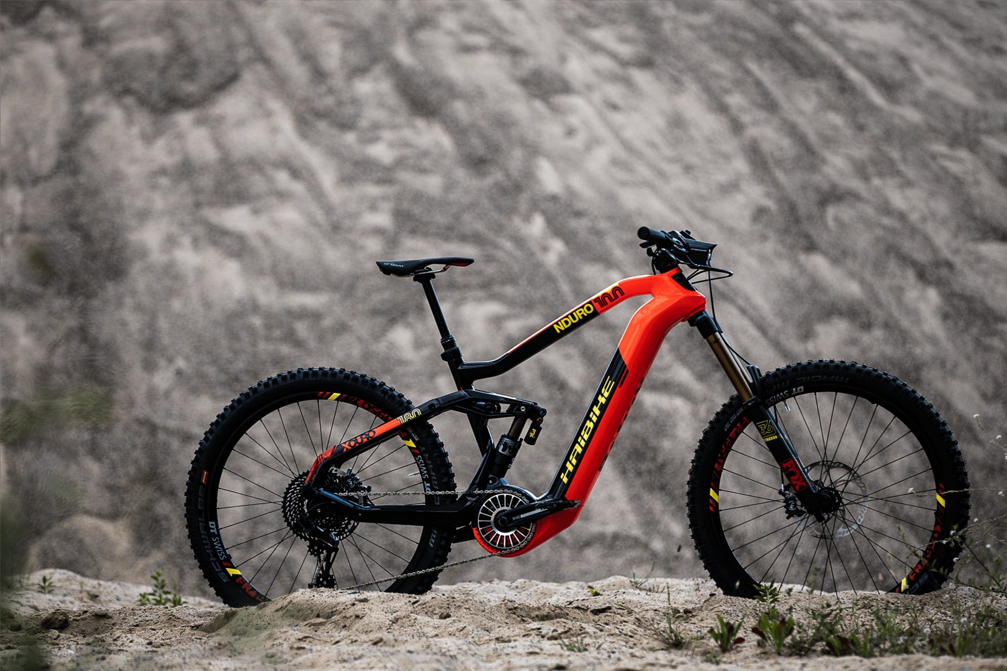 Ein modernes, orange-schwarzes E-Mountainbike steht dynamisch auf einem sandigen Untergrund. Der robuste Rahmen und