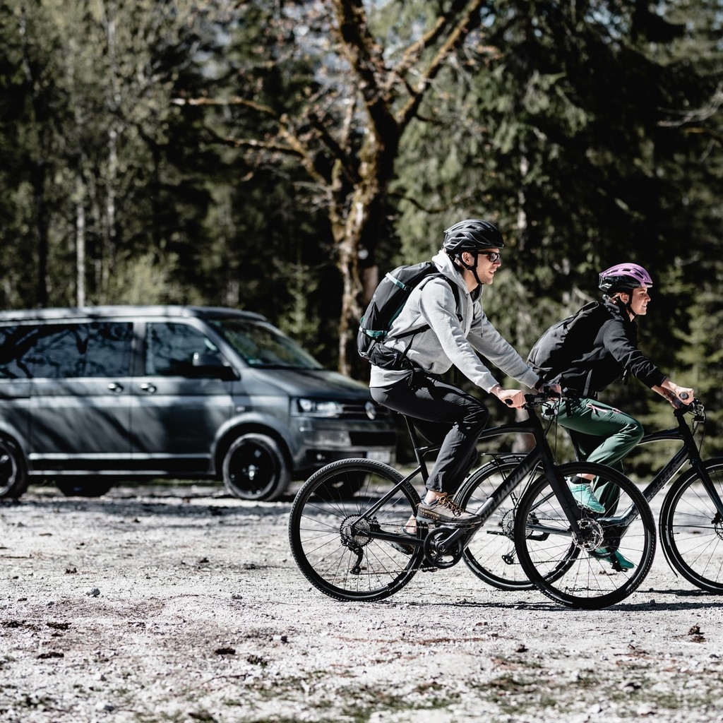 Zwei Personen fahren auf e-Bikes durch die Natur, im Hintergrund sieht man einen Transporter