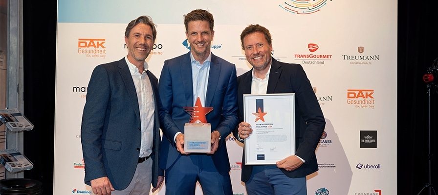 Drei Männer in Anzügen halten eine Trophäe und eine Urkunde bei einem Event mit Sponsoren-Logos im Hintergrund.